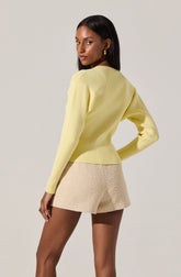 A woman is wearing a pastel yellow ribbed long-sleeve sweater paired with beige textured shorts. The sweater has a simple, fitted design with a round neckline, and the shorts are high-waisted with a slightly loose fit. She accessorizes with gold hoop earrings and a ring.